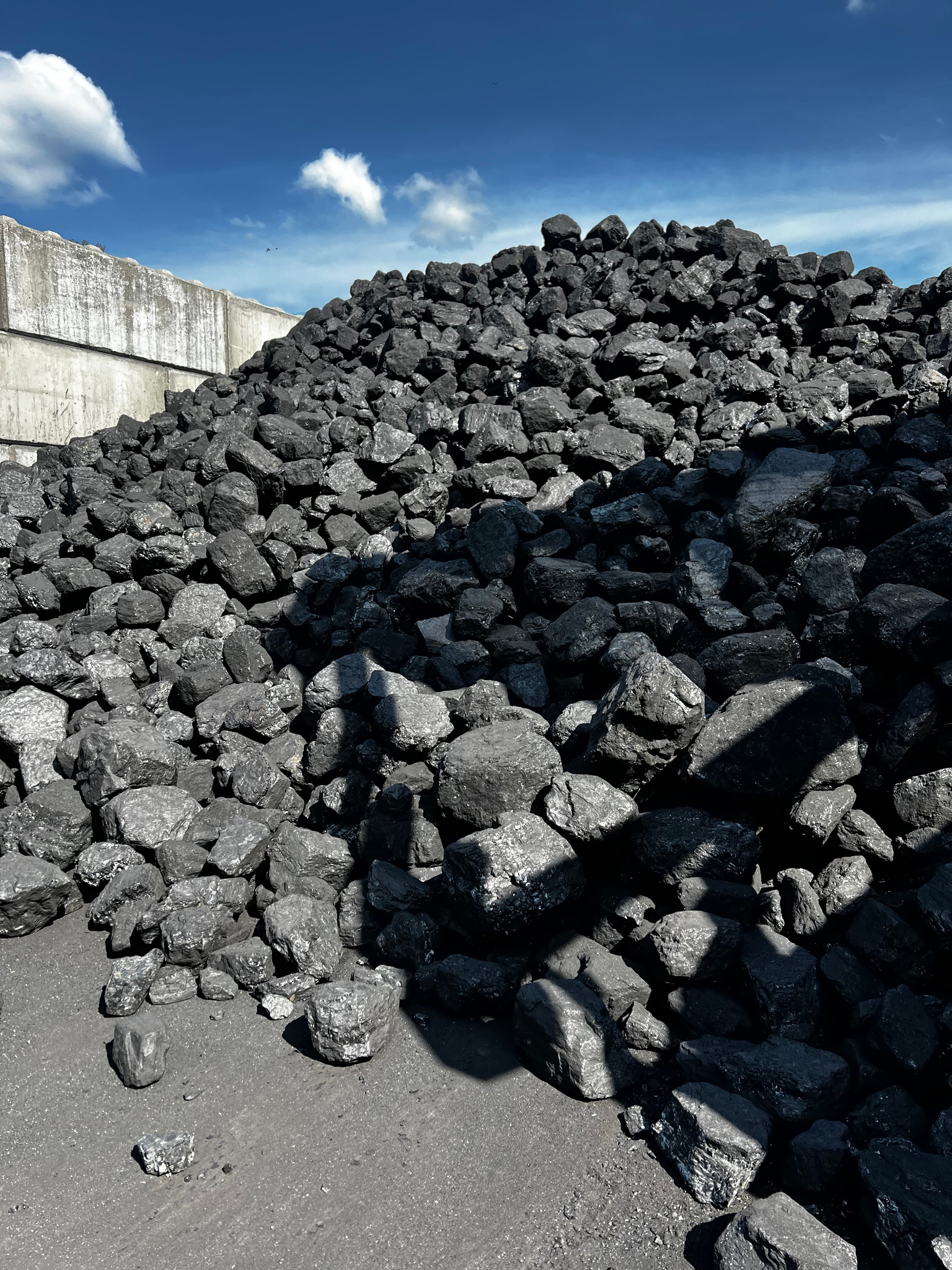 Góra czarnego węgla kamiennego pod błękitnym niebem z chmurami, obok betonowego muru. Widoczne ostre cienie i struktura materiału.