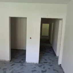 Wnętrze po tynkowaniu maszynowym i wylewce posadzki, widoczne otwory drzwiowe i surowe ściany. Szara posadzka z plamami.