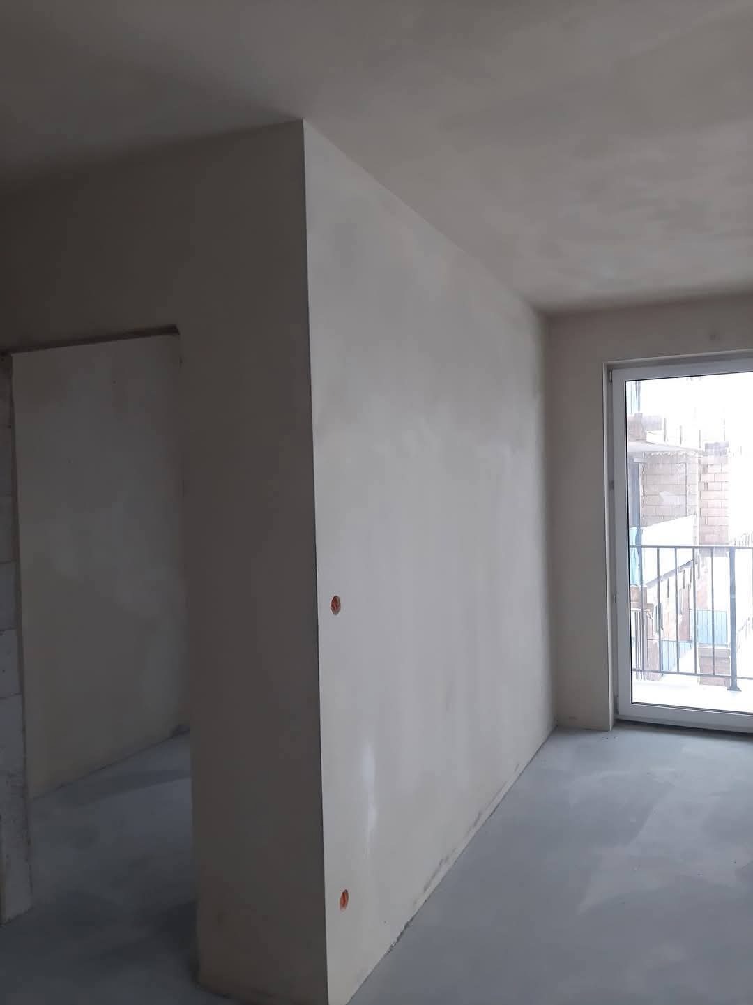 Wnętrze po tynkowaniu maszynowym, widoczne gładkie ściany i posadzka. Otwory na gniazdka elektryczne w ścianie. Okno balkonowe z widokiem na zewnątrz.