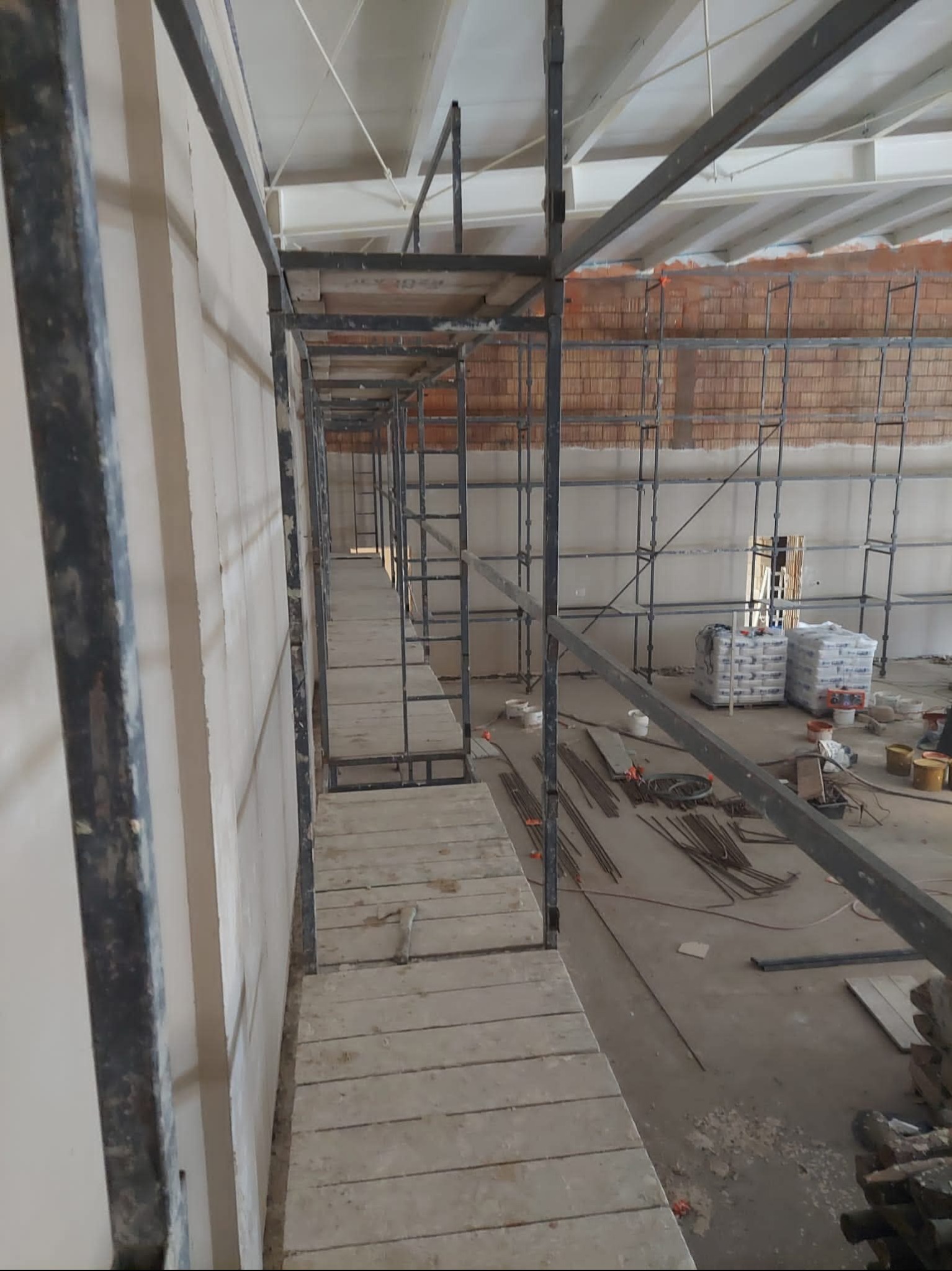 Wnętrze w trakcie prac tynkarskich: ściana po tynkowaniu, rusztowanie, materiały budowlane, narzędzia. Widok z perspektywy rusztowania, na dalszym planie ściana z cegły.
