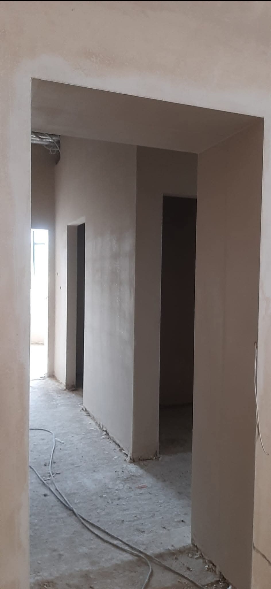 Wnętrze po tynkowaniu maszynowym: widok na korytarz z surową posadzką, ściany pokryte jasnym tynkiem, widoczne kable pod sufitem.