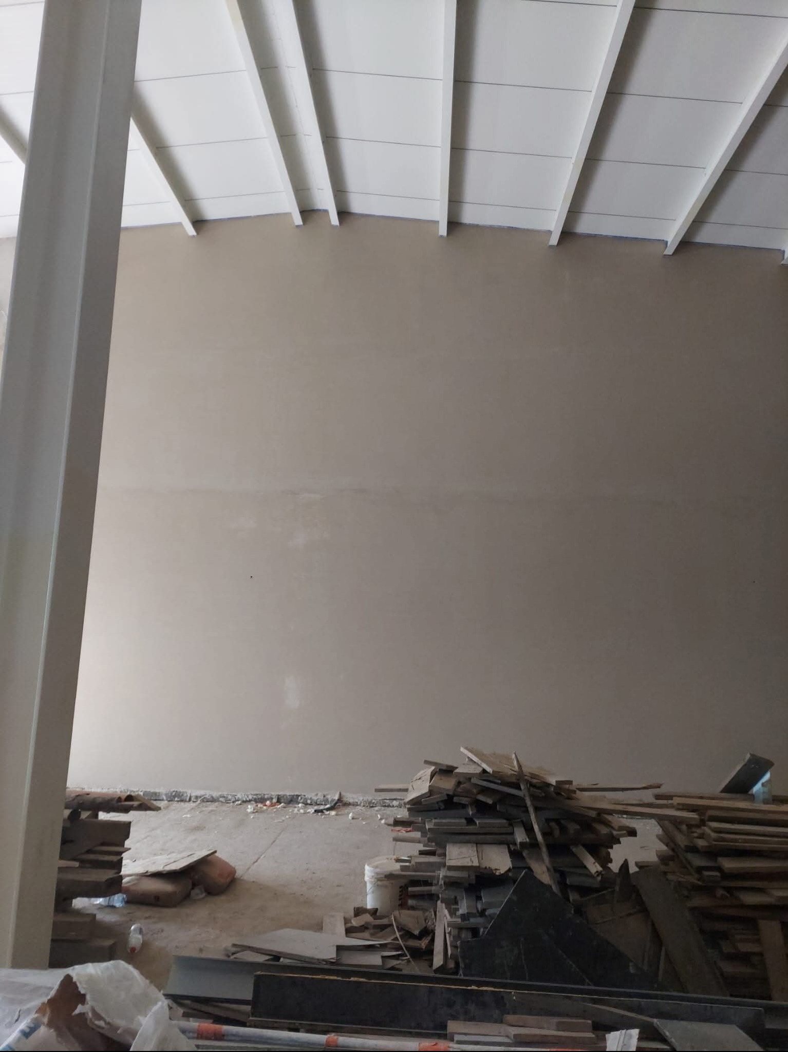 Świeżo otynkowana ściana w surowym wnętrzu z widoczną konstrukcją sufitu i stertą materiałów budowlanych na podłodze, ukazująca etap prac wykończeniowych.