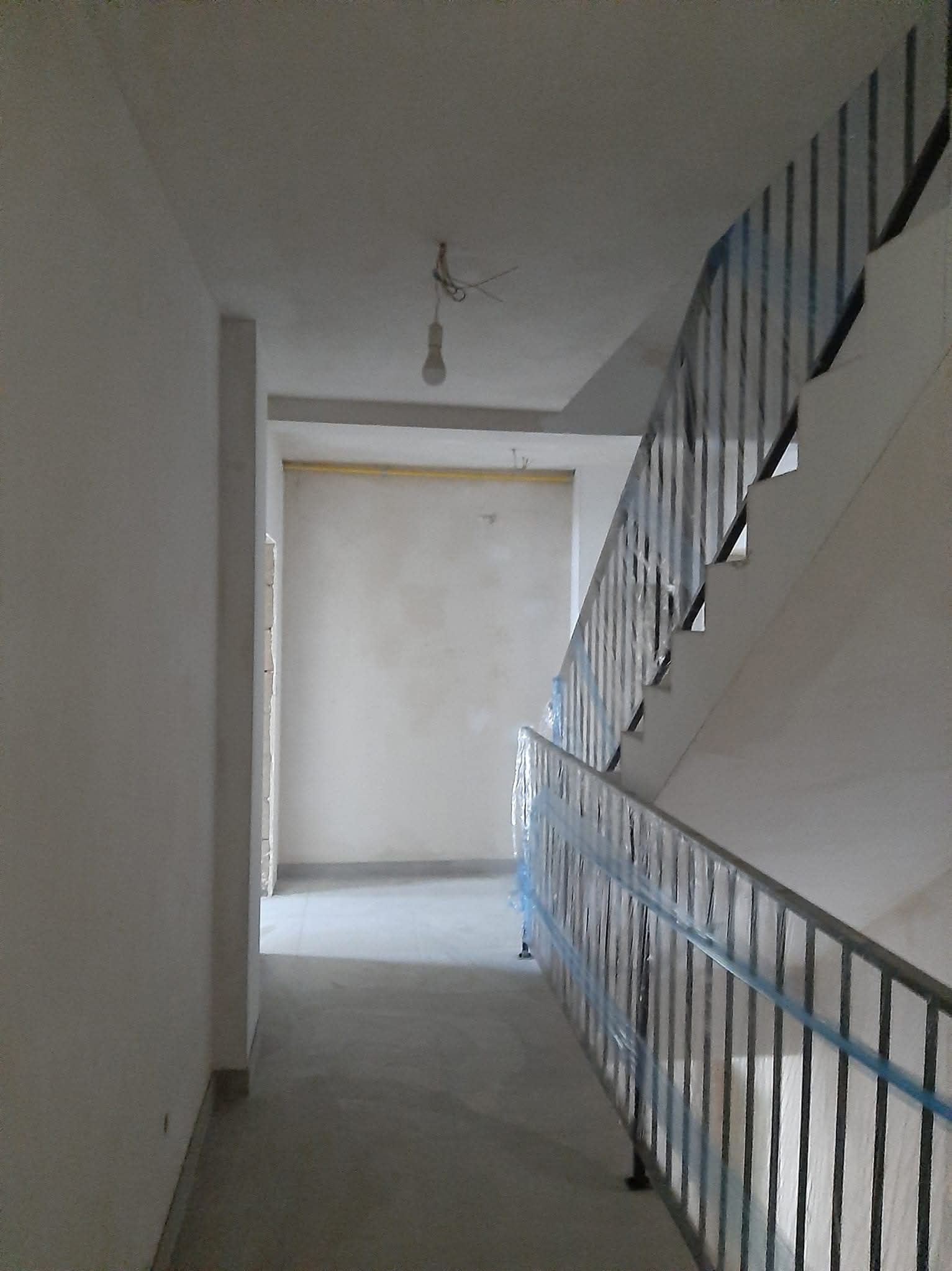 Klatka schodowa w trakcie prac wykończeniowych: ściany po tynkowaniu, posadzka wylewana, barierki schodowe zabezpieczone folią. Widok z dołu schodów.