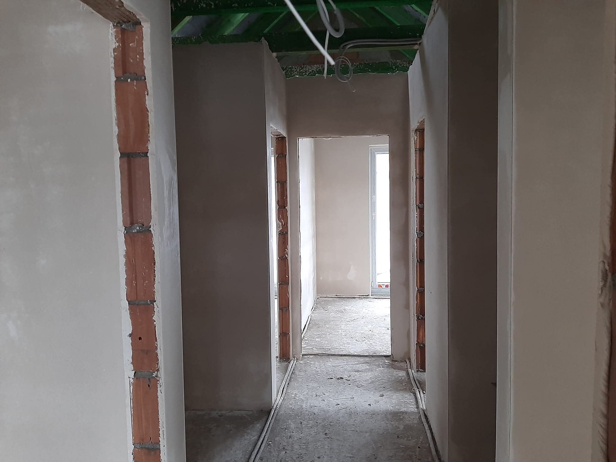 Wnętrze w trakcie tynkowania maszynowego: ściany częściowo pokryte tynkiem, widoczne fragmenty cegieł w otworach drzwiowych, surowa podłoga.