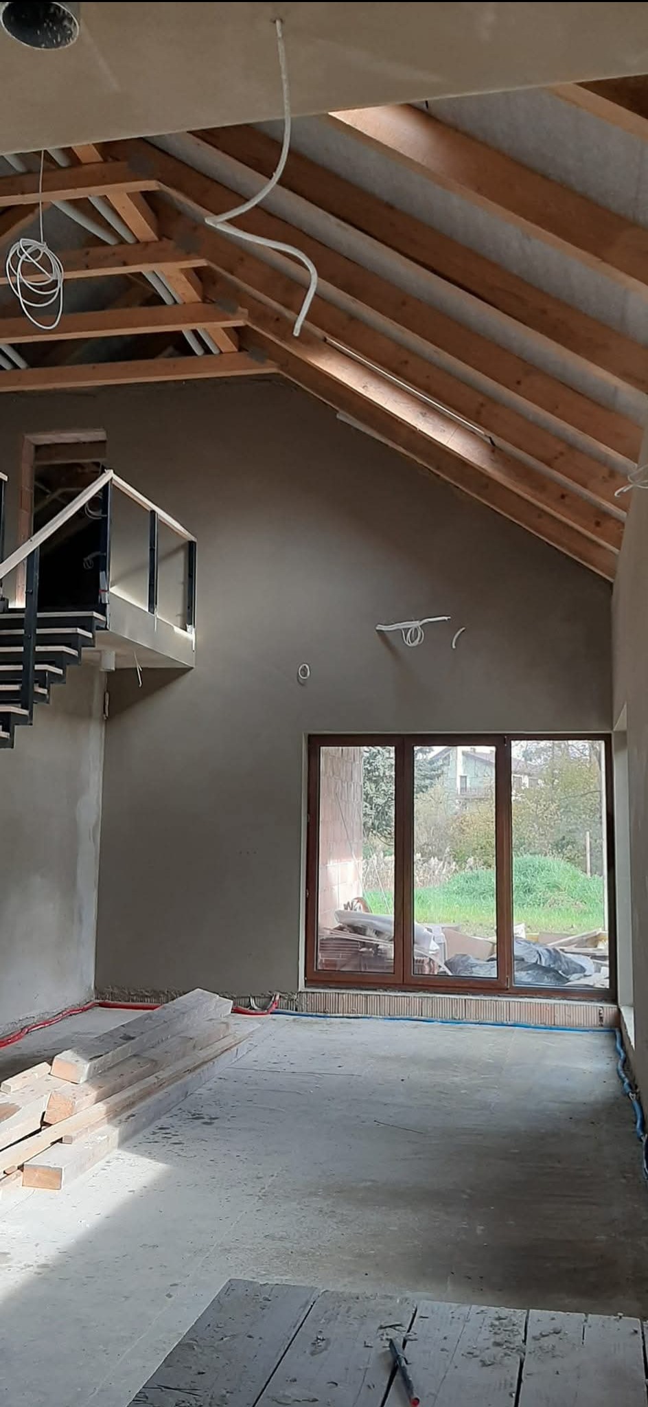 Wnętrze domu w stanie deweloperskim: surowe tynki na ścianach, drewniana konstrukcja dachu, schody z balustradą i okno z widokiem na zieleń. Widoczne instalacje.