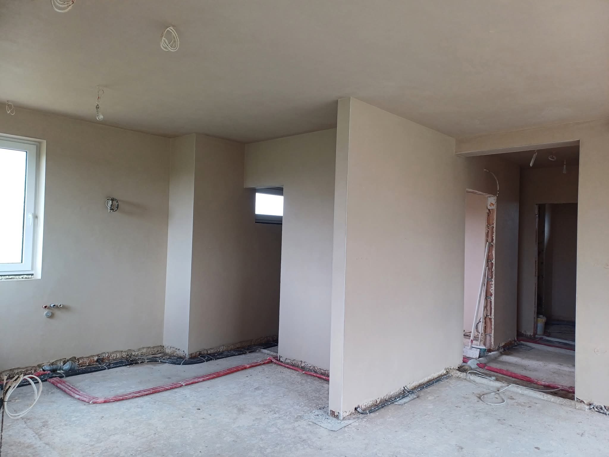 Wnętrze po tynkowaniu: gładkie ściany w beżowym odcieniu, widoczne instalacje elektryczne i hydrauliczne przed posadzką. Stan surowy, przygotowanie do dalszych prac wykończeniowych.