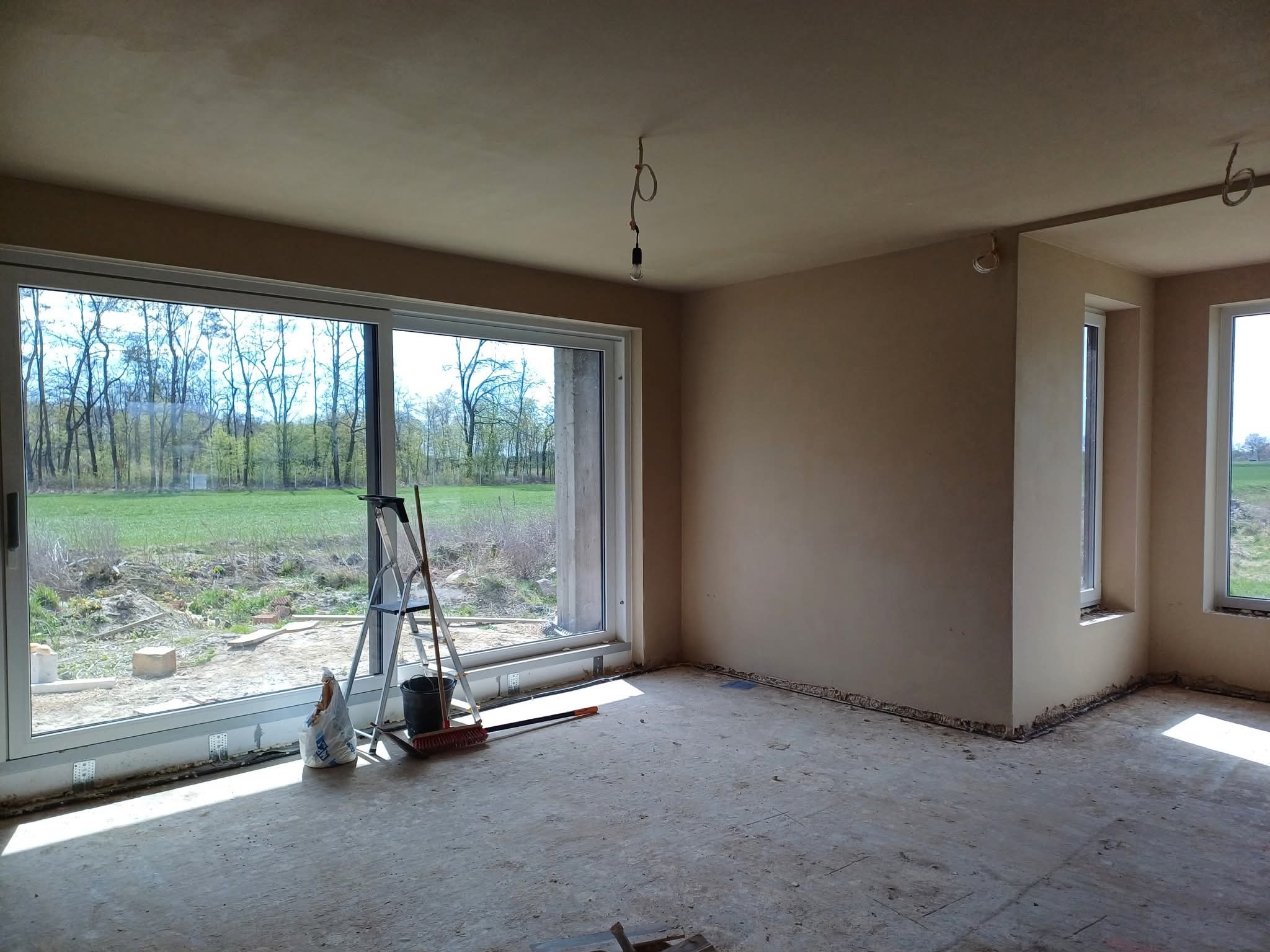 Wnętrze po tynkowaniu maszynowym: gładkie, beżowe ściany, okna z widokiem na zieleń, drabina i narzędzia na posadzce. Widoczne niedoskonałości posadzki przed wylaniem jastrychu.