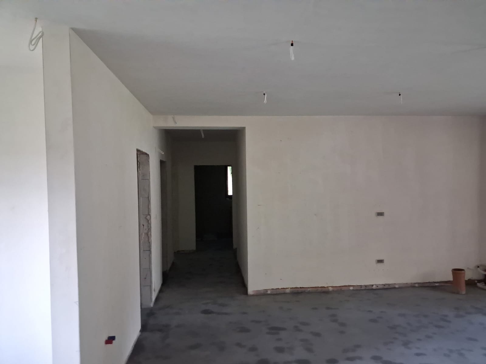 Wnętrze w stanie deweloperskim po tynkowaniu maszynowym i wylewce jastrychowej. Widoczne ściany z gładzią, wylewka na podłodze, wystające kable z sufitu.