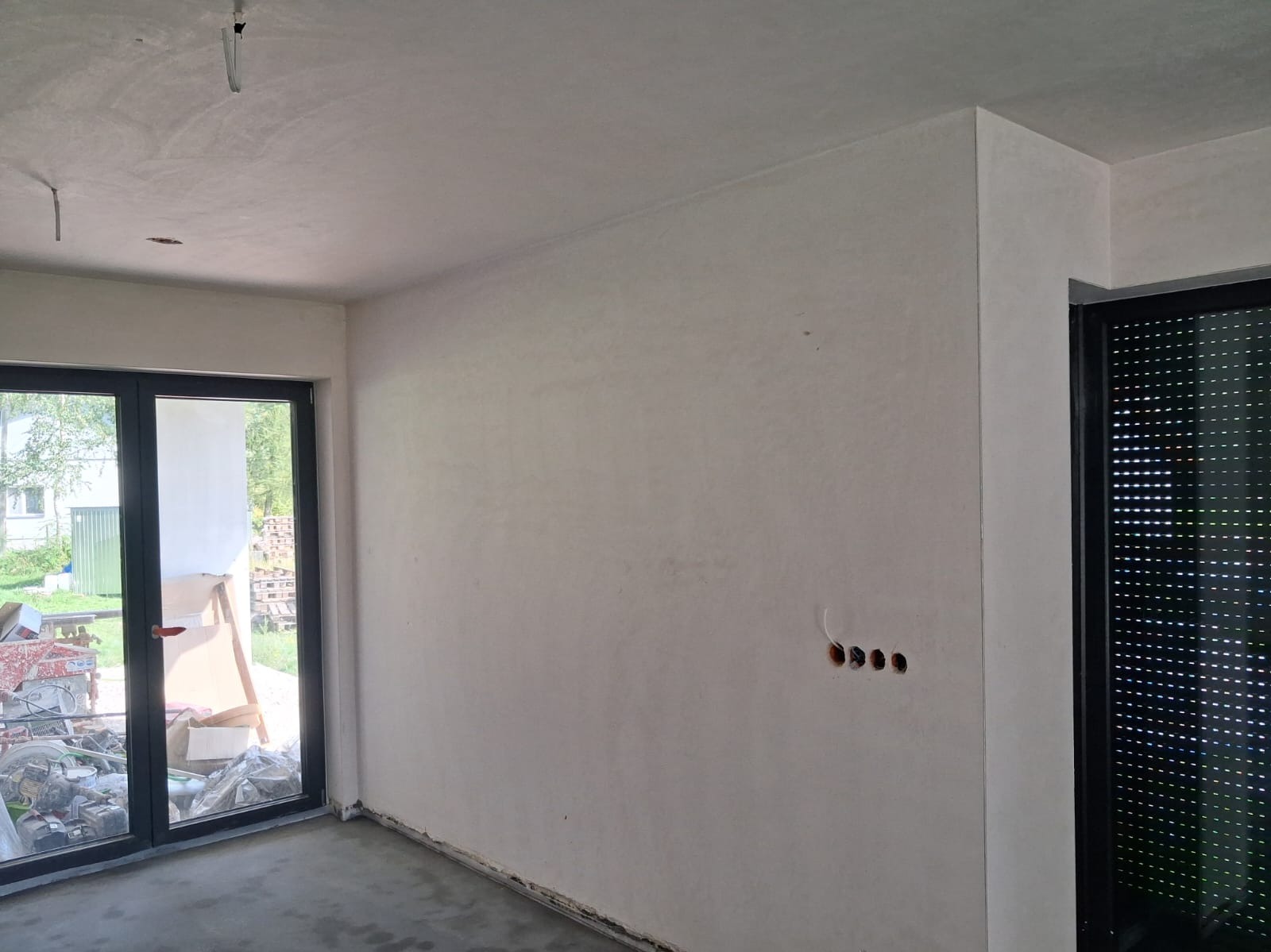 Wnętrze po tynkowaniu maszynowym: gładkie, jasne ściany w nowym domu, widoczne otwory na gniazdka elektryczne, okno z widokiem na zewnątrz i surowa posadzka.