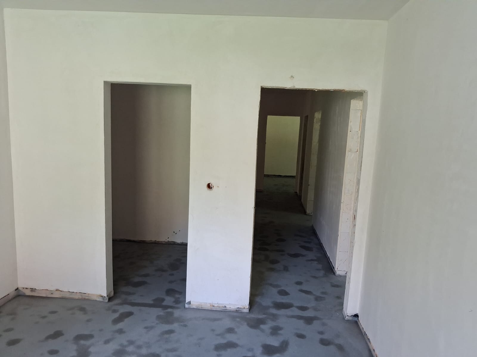 Wnętrze po tynkowaniu maszynowym i wylewce posadzki, widoczne otwory drzwiowe i surowe ściany. Szara posadzka z plamami.