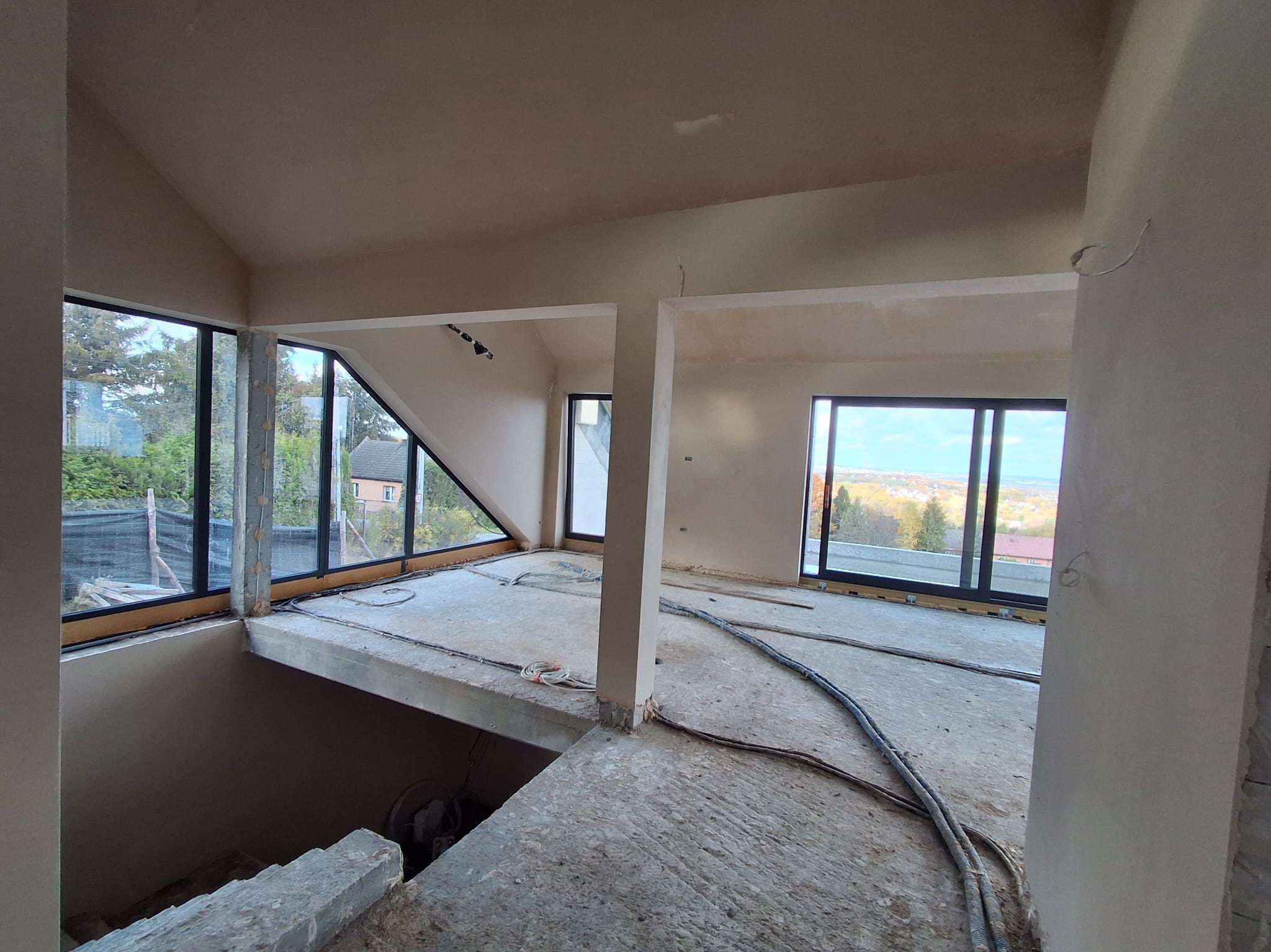 Wnętrze w trakcie prac wykończeniowych: świeżo otynkowane ściany i sufit, widoczne okna z czarnymi ramami, betonowa podłoga z przewodami. Surowy stan, przygotowanie do dalszych prac.