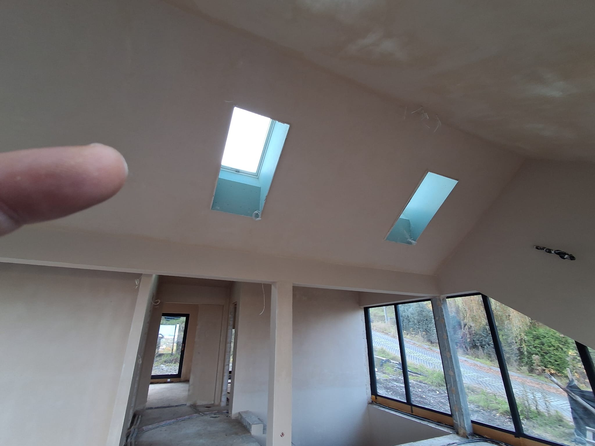 Wnętrze poddasza po tynkowaniu maszynowym, widoczne okna dachowe i ściany w jasnym kolorze. Na pierwszym planie fragment palca. Widok na zewnątrz przez okna.