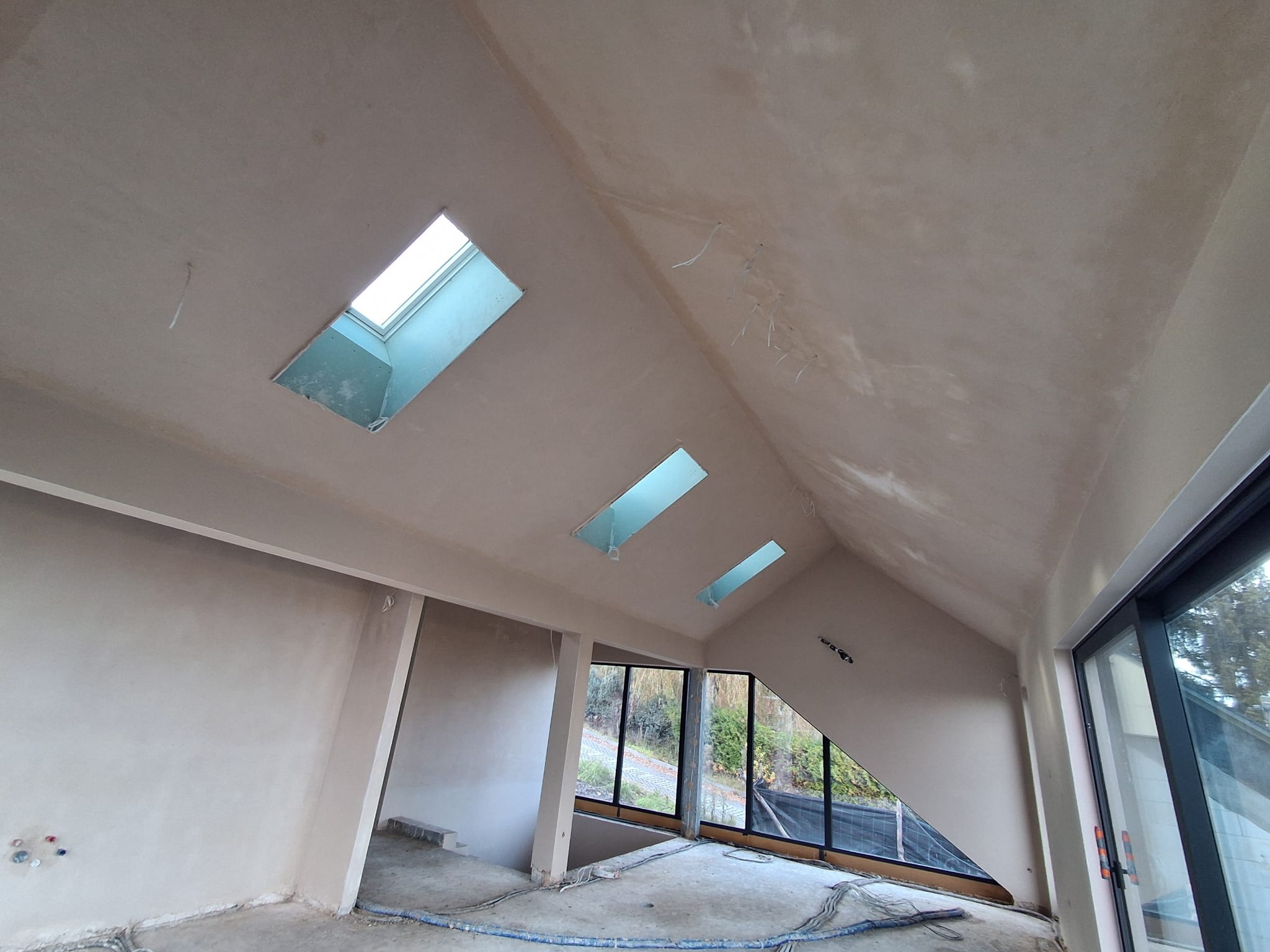 Wnętrze po tynkowaniu maszynowym w Tarnowie. Widoczne okna dachowe i duże przeszklenia. Ściany i sufit pokryte świeżym, jasnym tynkiem. Niebieskie węże na podłodze.