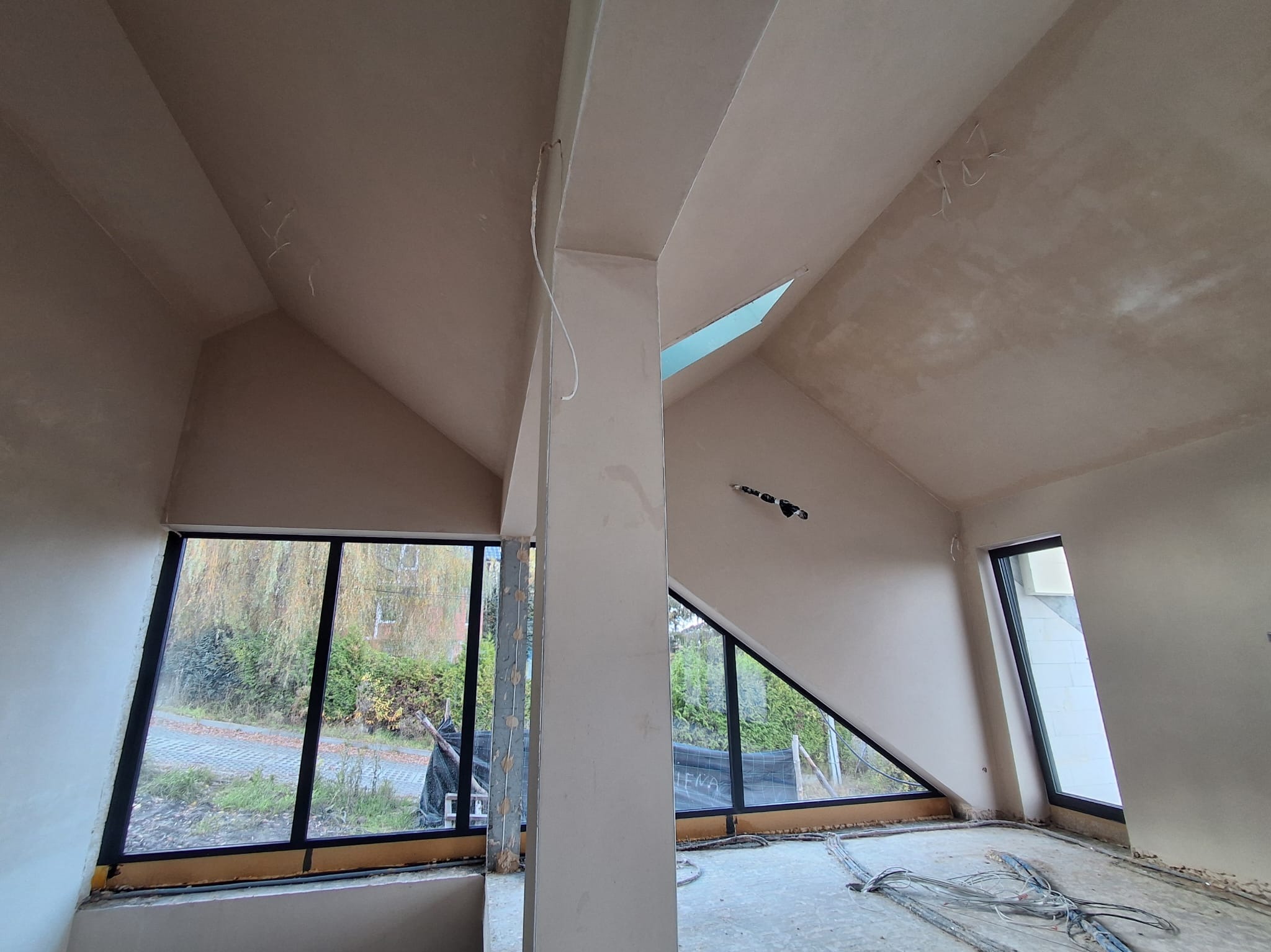 Wnętrze poddasza po tynkowaniu maszynowym, widoczne okna o nietypowych kształtach, surowe przewody elektryczne na podłodze, kolumna konstrukcyjna.