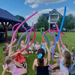 Animatorzy Niezapominajki - Grupa dzieci siedzi na trawie, trzymając kolorowe balony modelowane, pod okiem animatorki z kwiatem we włosach. W tle dom i altana.