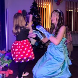 Animatorzy Niezapominajki - Animator w błękitnej sukni bawi się z dziewczynką w stroju Myszki Minnie na tle choinki i balonów. Uśmiechnięta animatorka i radosna atmosfera imprezy.