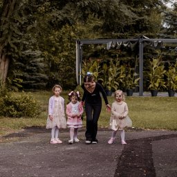 Animatorzy Niezapominajki - Animator maluje twarze dzieciom w parku, przygotowując je do zabawy. W tle altana i zieleń. Dziewczynki ubrane w sukienki, z pomalowanymi twarzami w motywy zwierzęce.