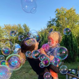 Animatorzy Niezapominajki - Animator dmucha bańki mydlane, trzymając dziecko z pomalowaną twarzą na tle zieleni i błękitnego nieba. Kolorowe bańki unoszą się w powietrzu.