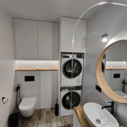 Nowoczesna łazienka z zabudowaną pralką i suszarką, umywalką na blacie drewnianym i okrągłym lustrem z podświetleniem LED. Minimalistyczny design, białe płytki, drewnopodobna podłoga.