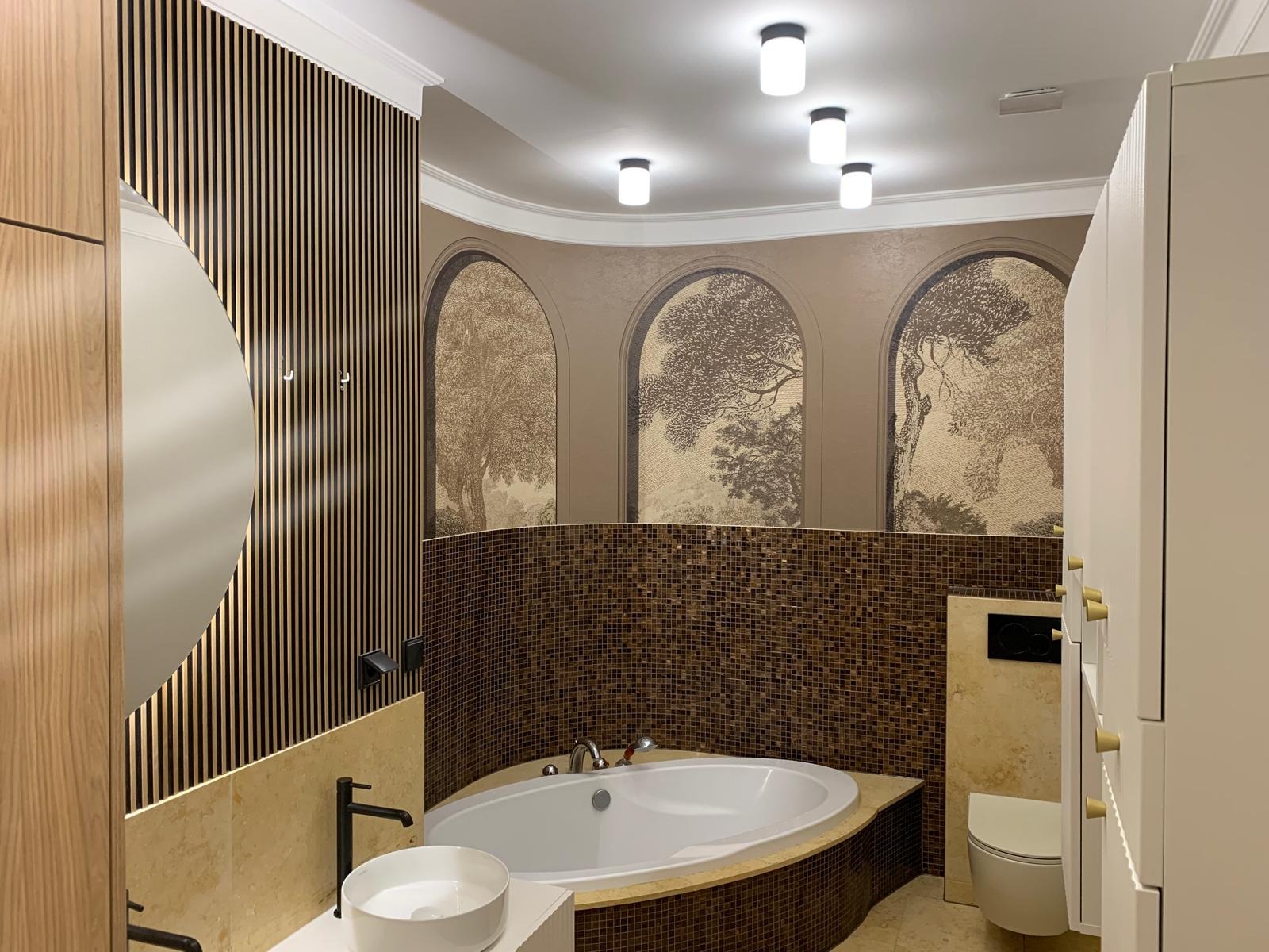 Elegancka łazienka z mozaiką i drewnianymi lamelami. Uwagę przykuwają grafiki drzew w niszach. Nowoczesna armatura i oświetlenie dopełniają aranżację.