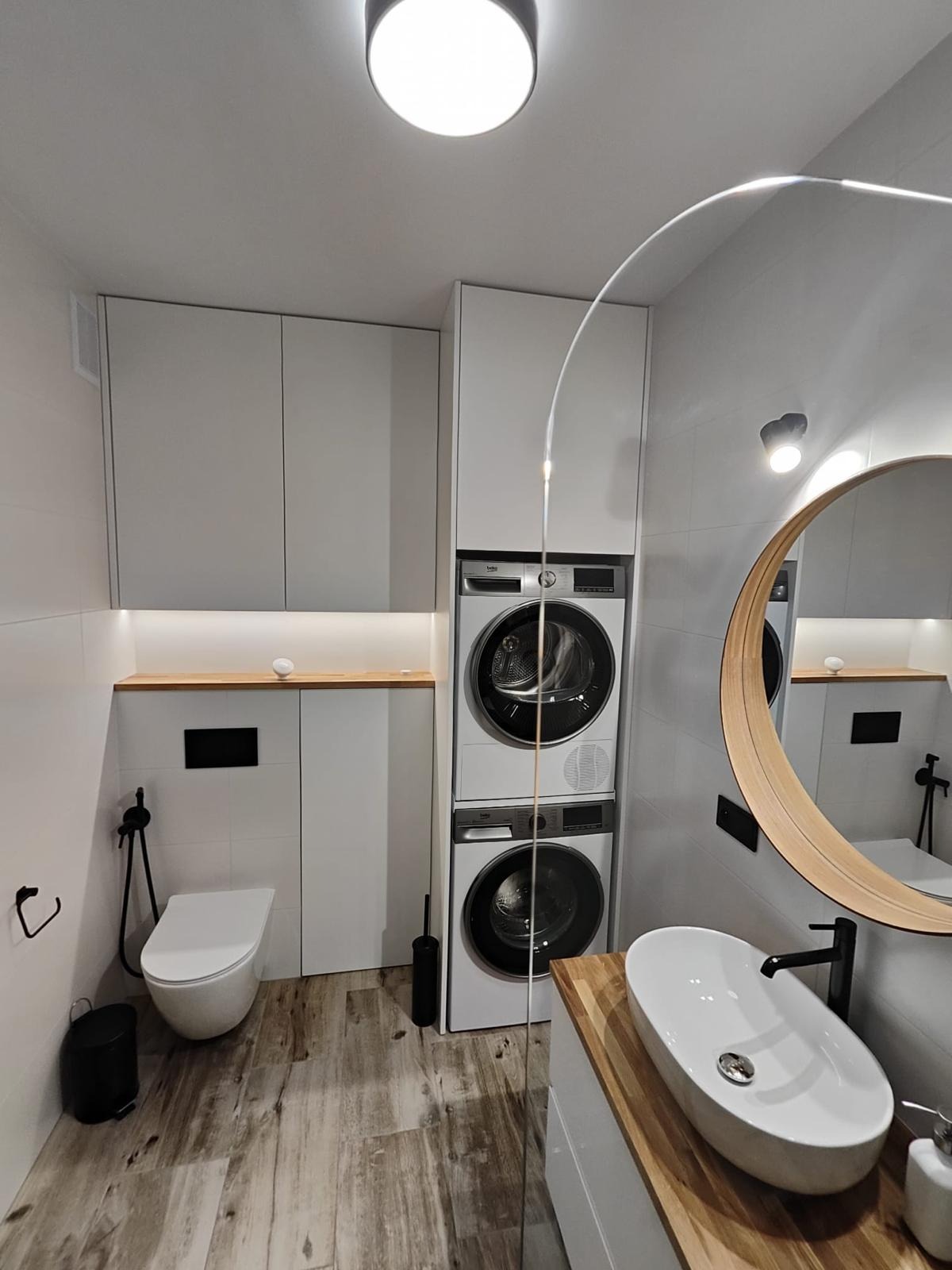 Nowoczesna łazienka z zabudowaną pralką i suszarką, umywalką na blacie drewnianym i okrągłym lustrem z podświetleniem LED. Minimalistyczny design, białe płytki, drewnopodobna podłoga.