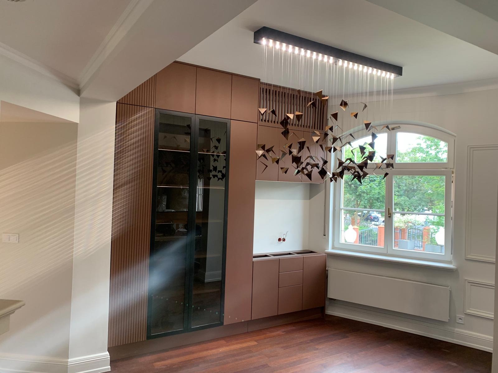 Nowoczesna zabudowa kuchenna w odcieniach brązu z przeszklonymi witrynami i oryginalną lampą wiszącą. Widok na zewnątrz przez okno.