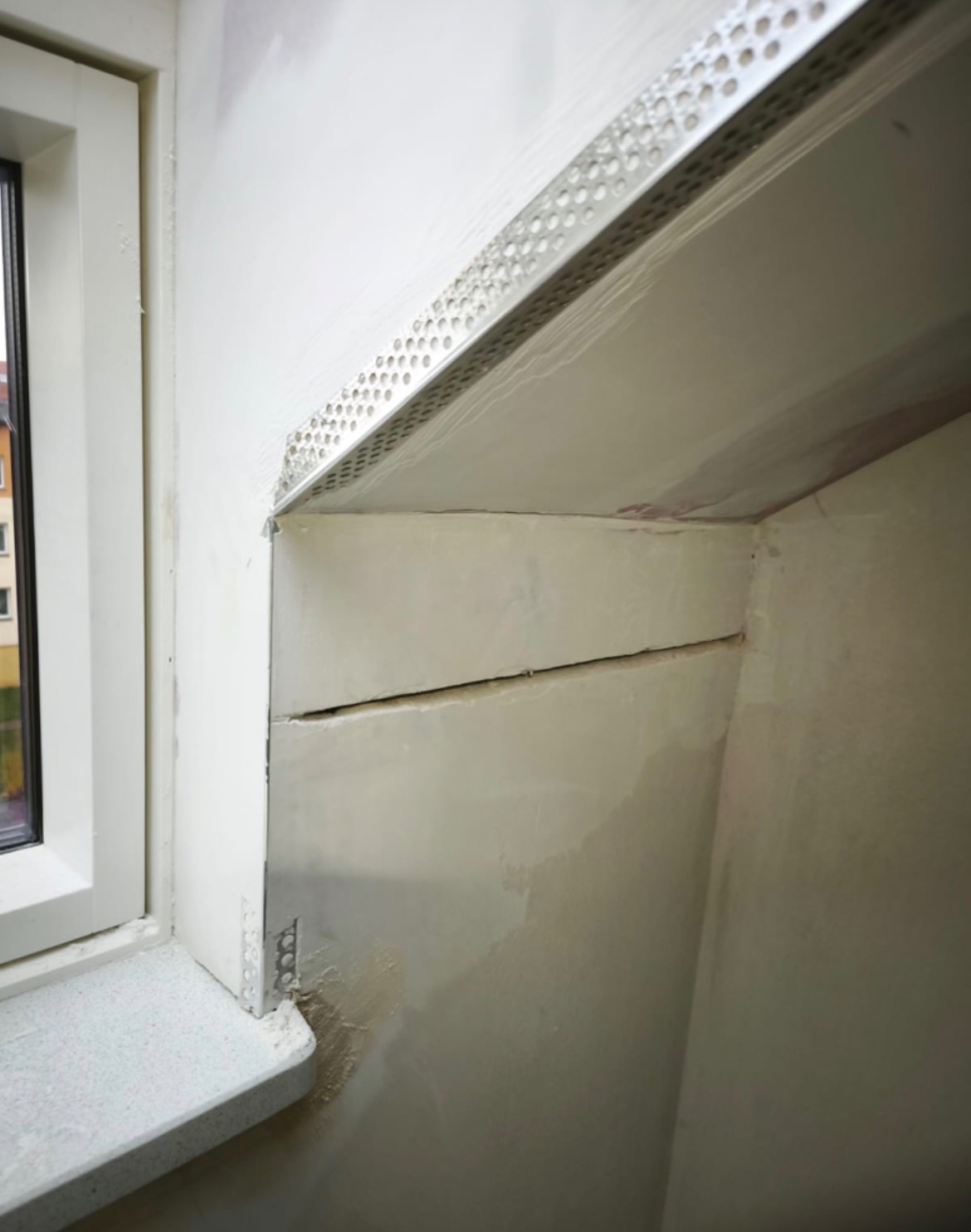 Narożnik ściany po szpachlowaniu, z widoczną listwą narożnikową i fragmentem okna. Ściana przygotowana do dalszych prac wykończeniowych, widoczne niedoskonałości.