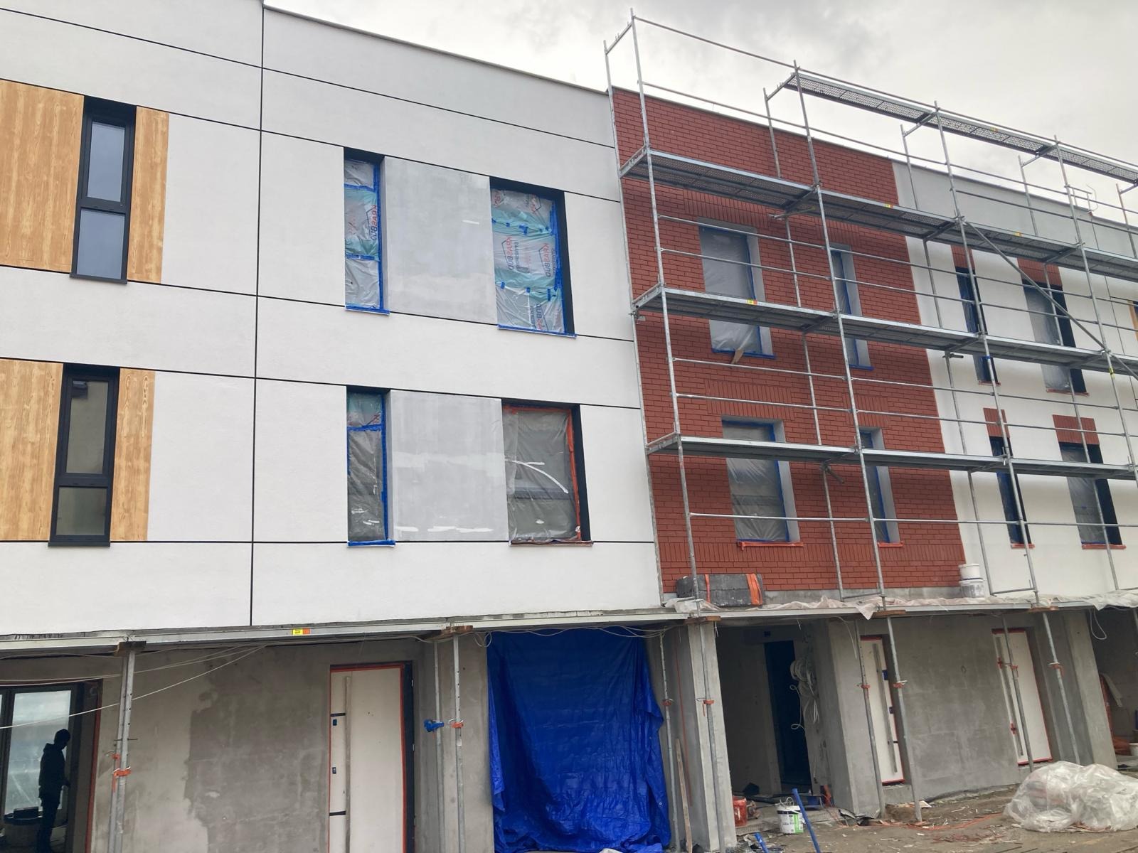 Budynek w trakcie wykończenia elewacji: część biała z drewnianymi akcentami, część z cegły, okna zabezpieczone folią, rusztowania. Prace budowlane w toku.