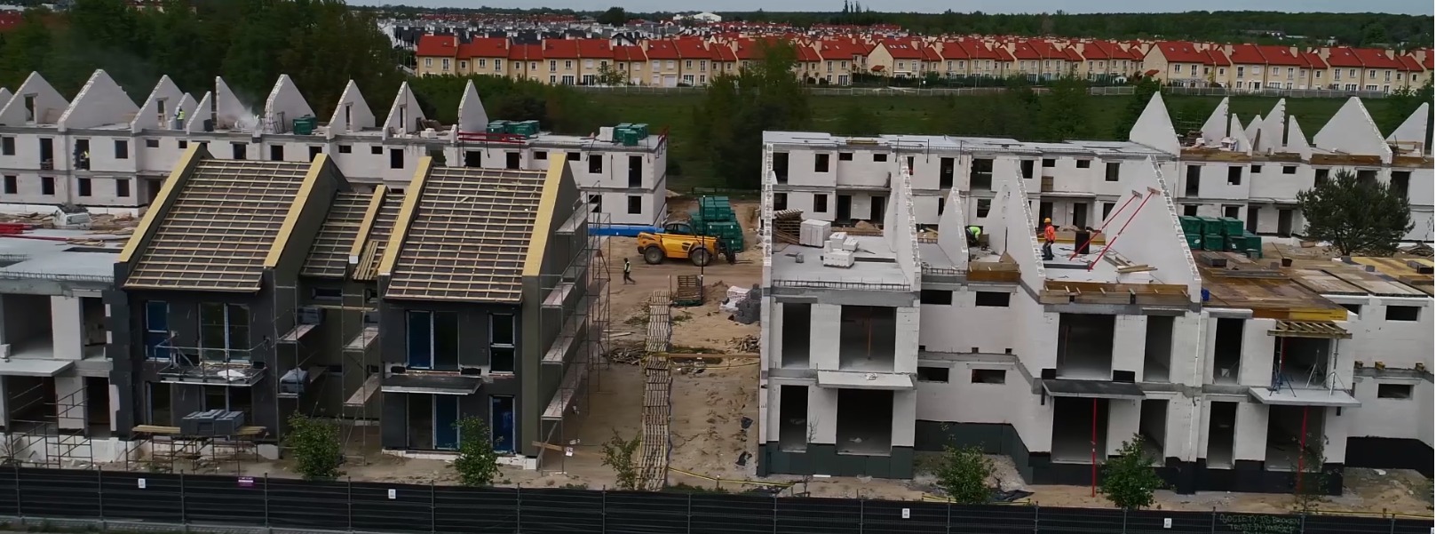 Osiedle w budowie z bliska: domy w różnym stadium, widoczne rusztowania, materiały budowlane i pracujący robotnicy. W tle rząd identycznych budynków.