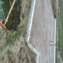 DOM PROJEKT SPÓŁKA Z OGRANICZONĄ ODPOWIEDZIALNOŚCIĄ - Etap budowy fundamentów: wykop z izolacją, widoczne szalunki i rury odprowadzające wodę. Teren błotnisty, częściowo zalany wodą. W tle zieleń.