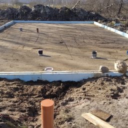 DOM PROJEKT SPÓŁKA Z OGRANICZONĄ ODPOWIEDZIALNOŚCIĄ - Wyrównany teren pod budowę domu, obramowany styropianem. Pracownik w żółtej kamizelce zagęszcza podłoże ubijarką. Widoczne wystające rury kanalizacyjne.