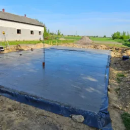 Świeżo wylana, gładka płyta fundamentowa z hydroizolacją, widok z góry. W tle budynek w stanie surowym otwartym i sprzęt geodezyjny. Błękitne niebo.