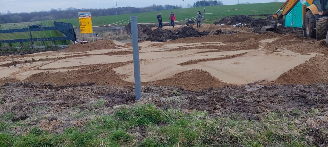 Przygotowany teren pod fundamenty z widoczną rurą kanalizacyjną. W tle pracownicy i maszyna budowlana. Teren wyrównany i przygotowany do dalszych prac.