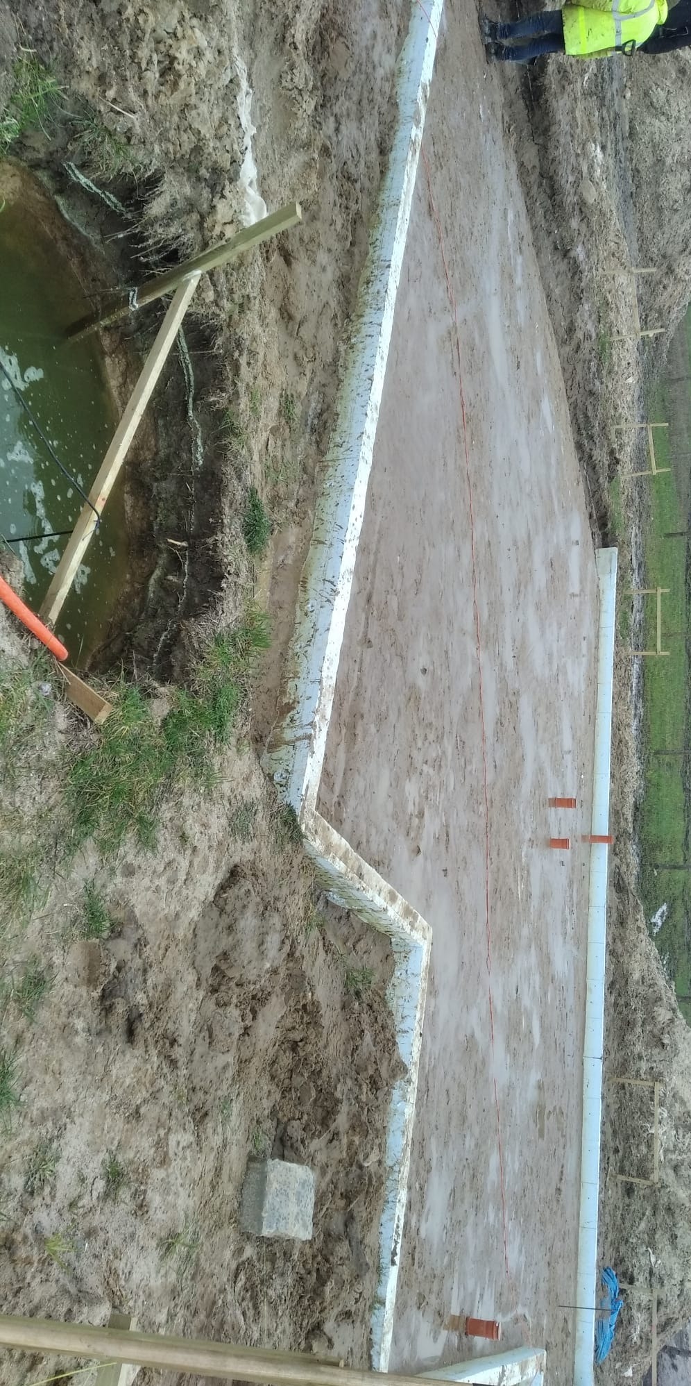 Etap budowy fundamentów: wykop z izolacją, widoczne szalunki i rury odprowadzające wodę. Teren błotnisty, częściowo zalany wodą. W tle zieleń.