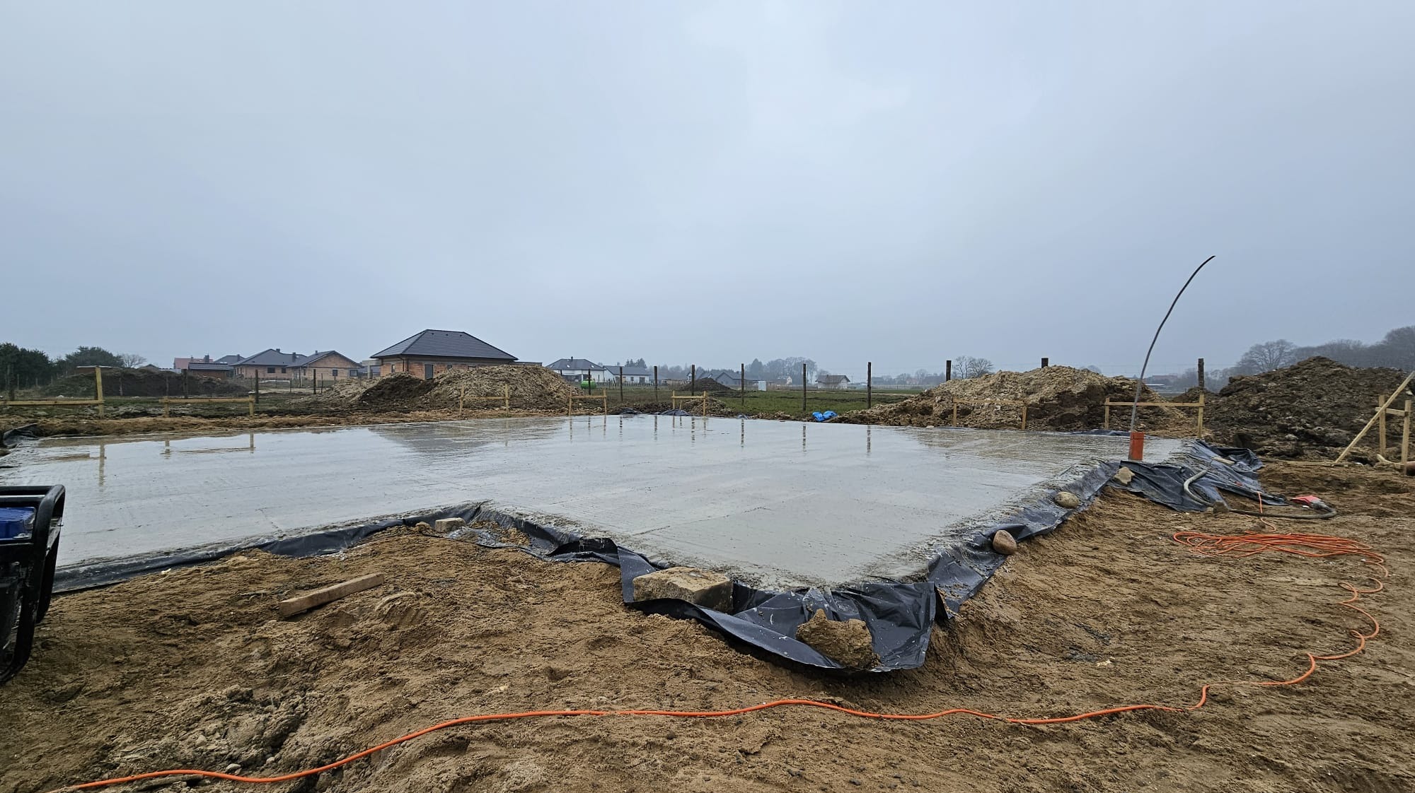 Zalany fundament pod dom jednorodzinny, zabezpieczony folią. Widoczne przygotowane wykopy i drewniane szalunki. W tle zabudowa jednorodzinna.