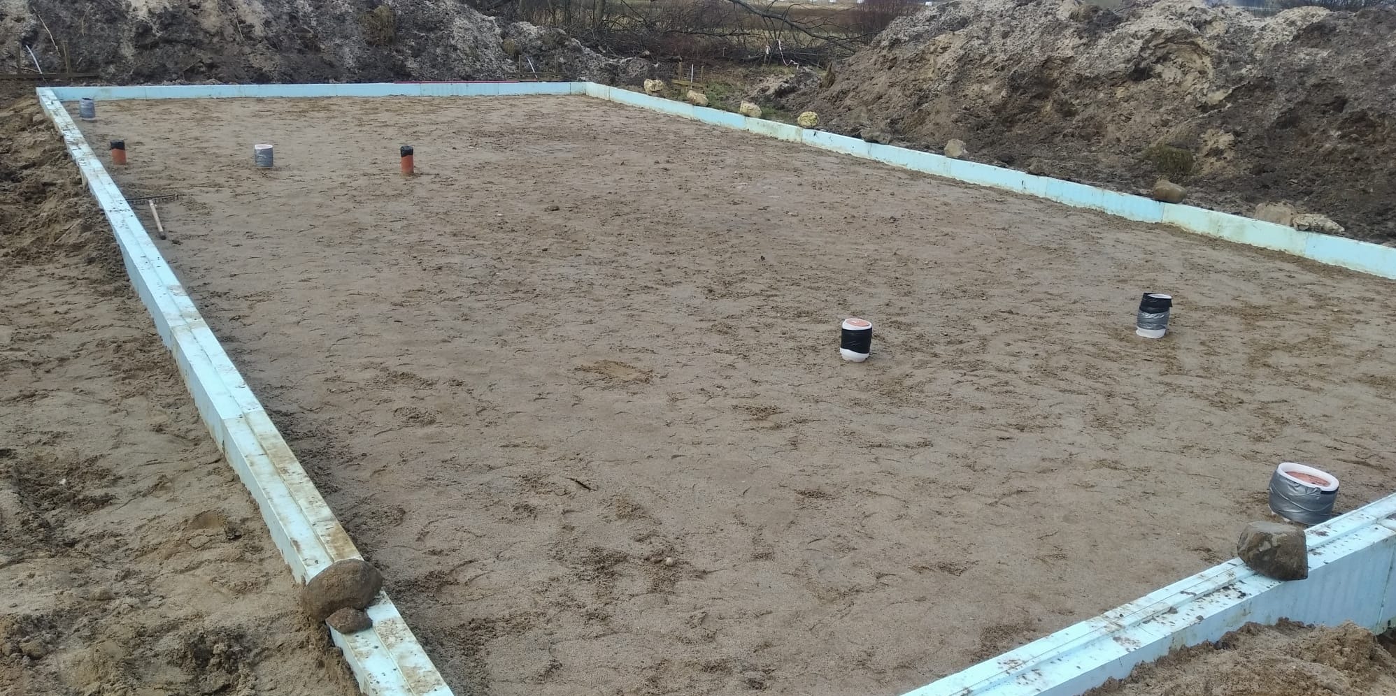 Wykop pod fundamenty domu jednorodzinnego, obłożony izolacją termiczną, z widocznymi pionami geodezyjnymi. Tło stanowią pryzmy ziemi i drzewa.