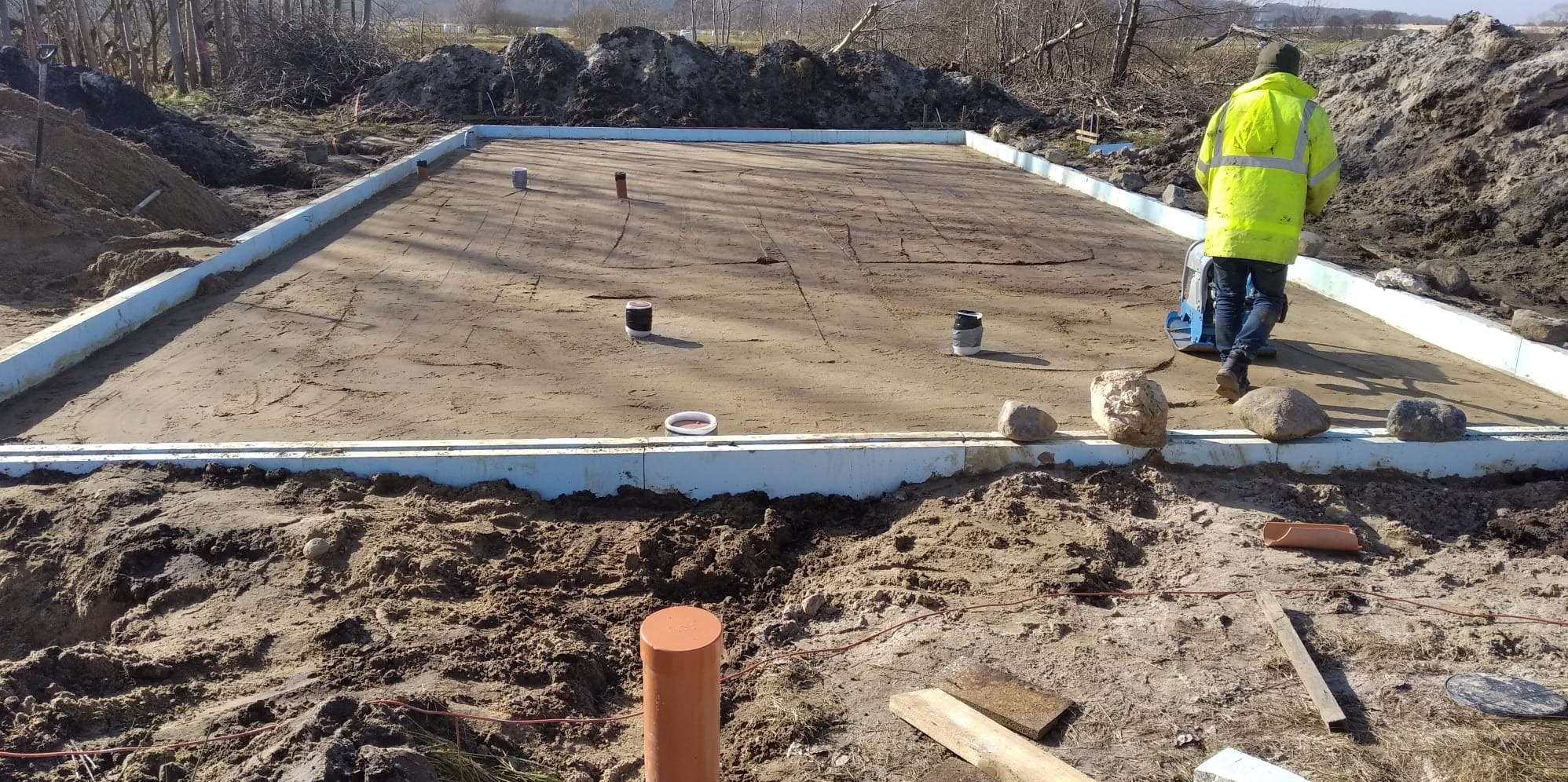 Wyrównany teren pod budowę domu, obramowany styropianem. Pracownik w żółtej kamizelce zagęszcza podłoże ubijarką. Widoczne wystające rury kanalizacyjne.