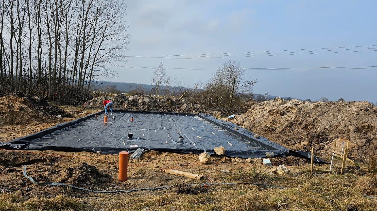 Przygotowanie terenu pod fundament: wykop zabezpieczony folią, widoczne elementy instalacji, osoba w tle, ziemia i drzewa. Etap prac ziemnych.