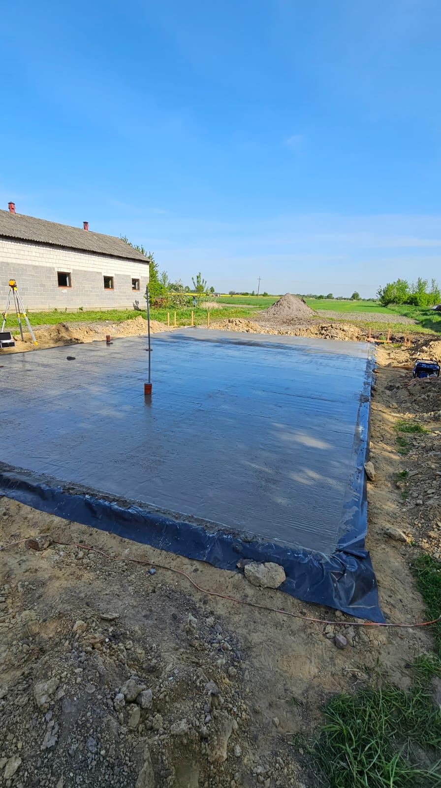 Świeżo wylana, gładka płyta fundamentowa z hydroizolacją, widok z góry. W tle budynek w stanie surowym otwartym i sprzęt geodezyjny. Błękitne niebo.