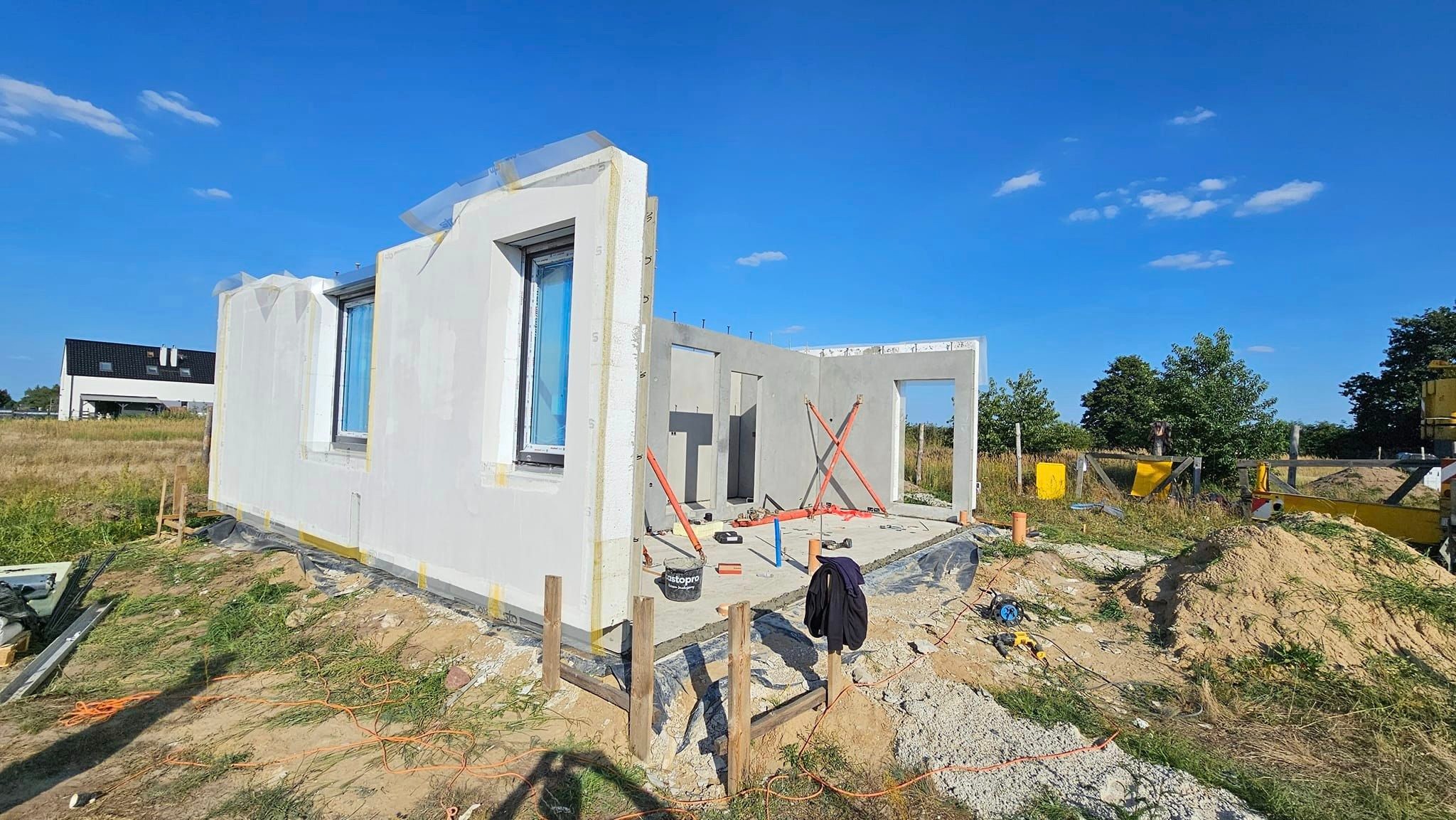 Wznoszenie domu z prefabrykatów: widoczne ściany z oknami, wsparte pomarańczowymi stemplami, na fundamencie. Na zewnątrz narzędzia i sprzęt budowlany.