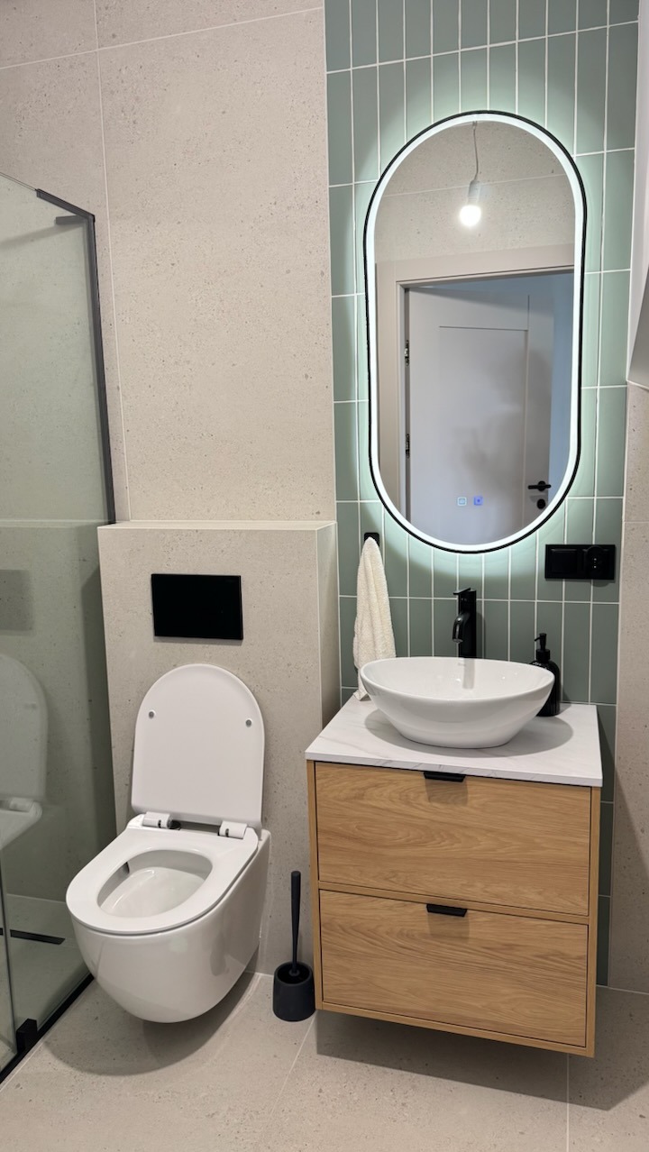 Nowoczesna łazienka z podwieszaną toaletą, umywalką nablatową na drewnianej szafce i podświetlanym lustrem w kształcie łuku. Jasne płytki i minimalistyczny design.