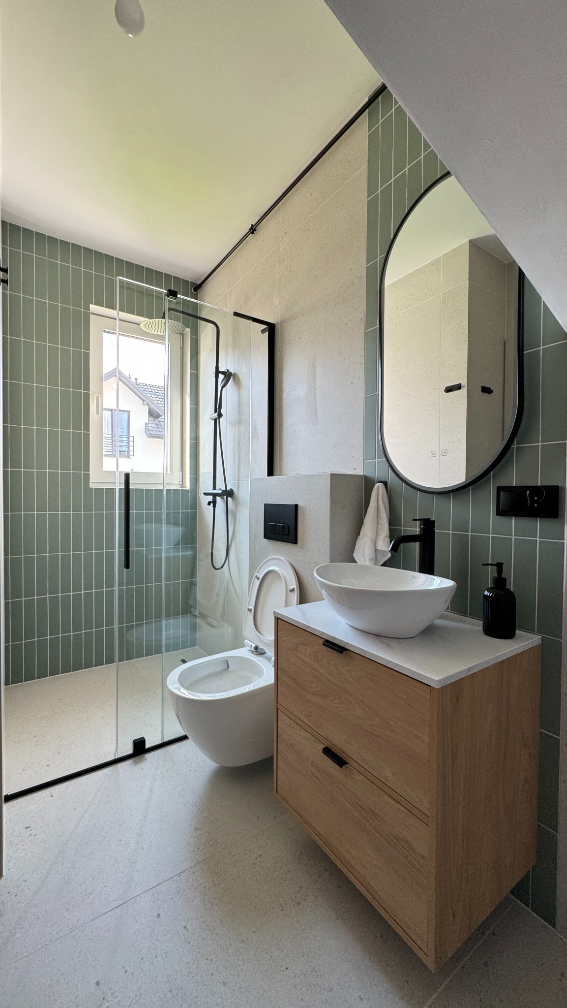 Nowoczesna łazienka z zielonymi płytkami, kabiną prysznicową typu walk-in, wiszącą toaletą i umywalką nablatową na drewnianej szafce. Minimalistyczny design z czarnymi akcentami.