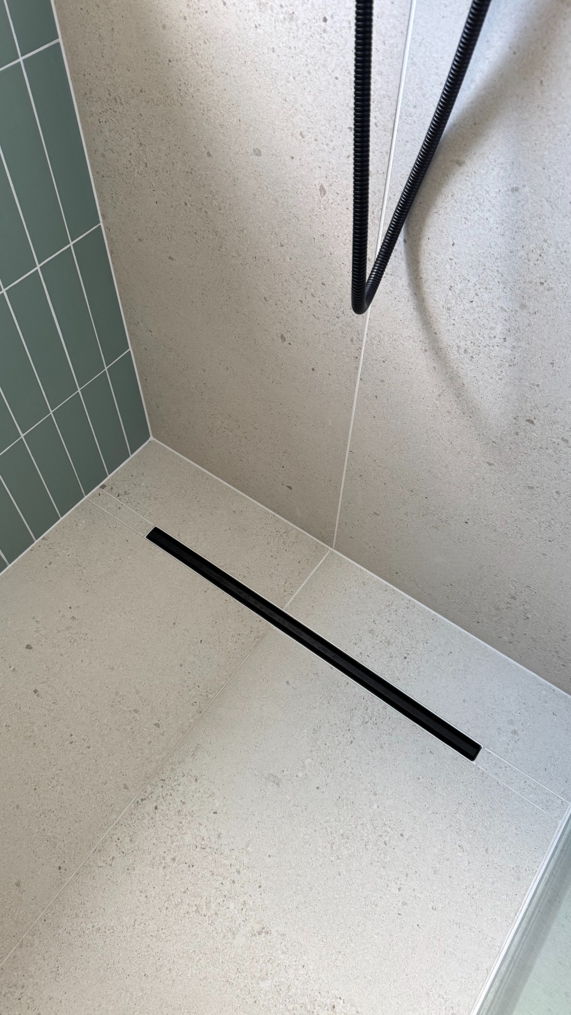 Wykończona strefa prysznicowa z odpływem liniowym w posadzce z jasnych płytek. Na ścianie zielone, pionowe płytki. Widoczny fragment węża prysznicowego.