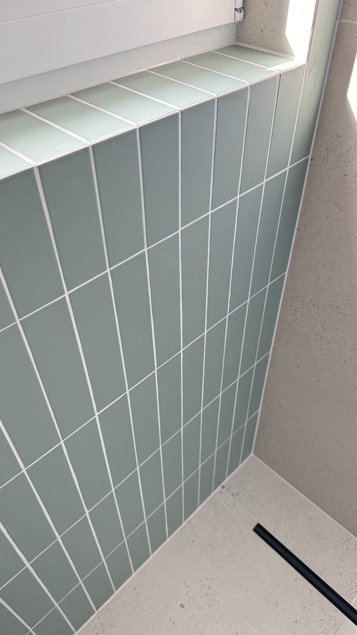 Ściana wyłożona pionowymi, zielonymi płytkami z białymi fugami, widoczny fragment podłogi z odpływem liniowym. Minimalistyczne wykończenie łazienki.