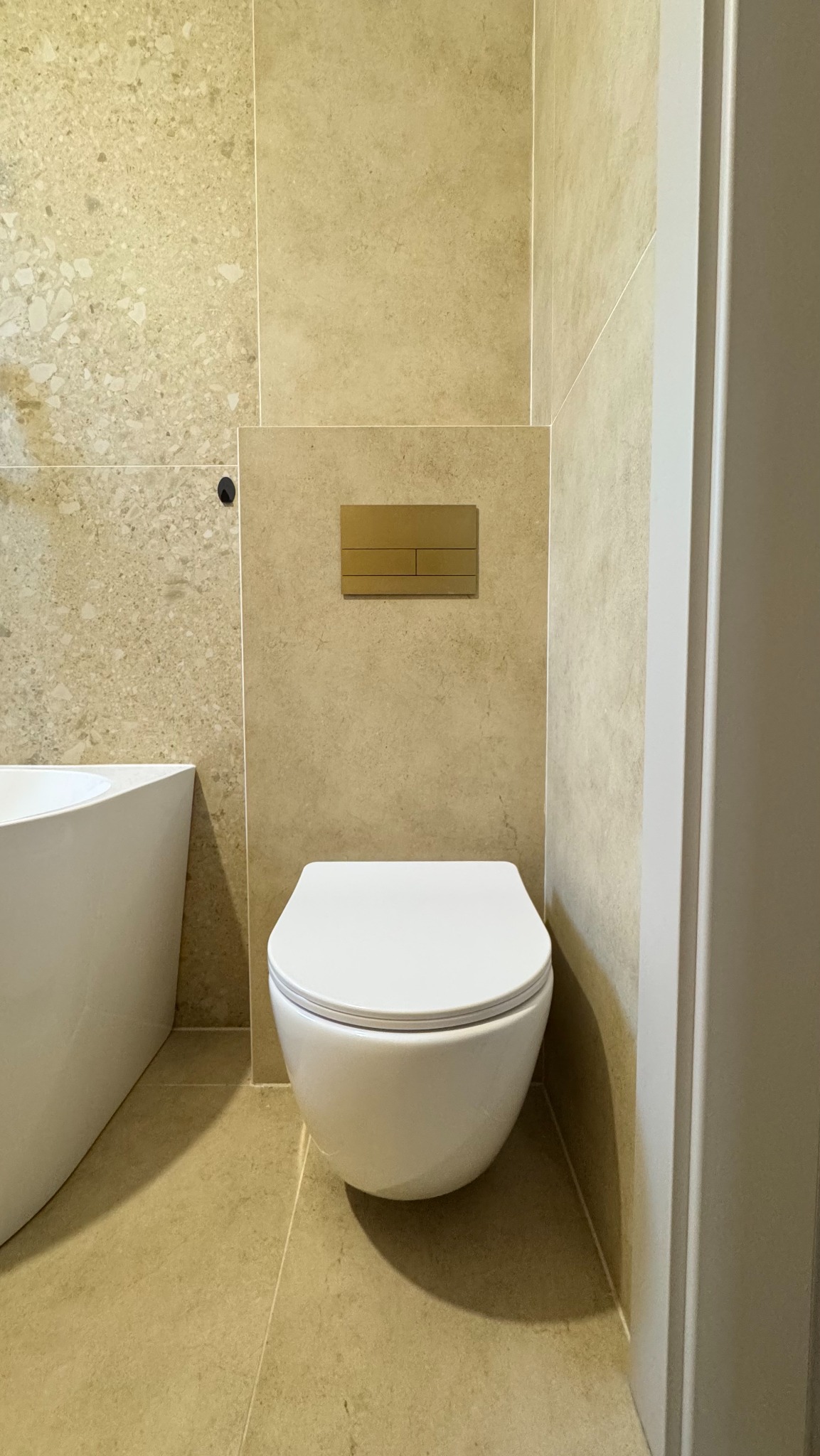 Nowoczesna łazienka po remoncie: podwieszana toaleta z beżowymi płytkami i złotym przyciskiem spłukującym. Minimalistyczny design, widoczny fragment wanny.