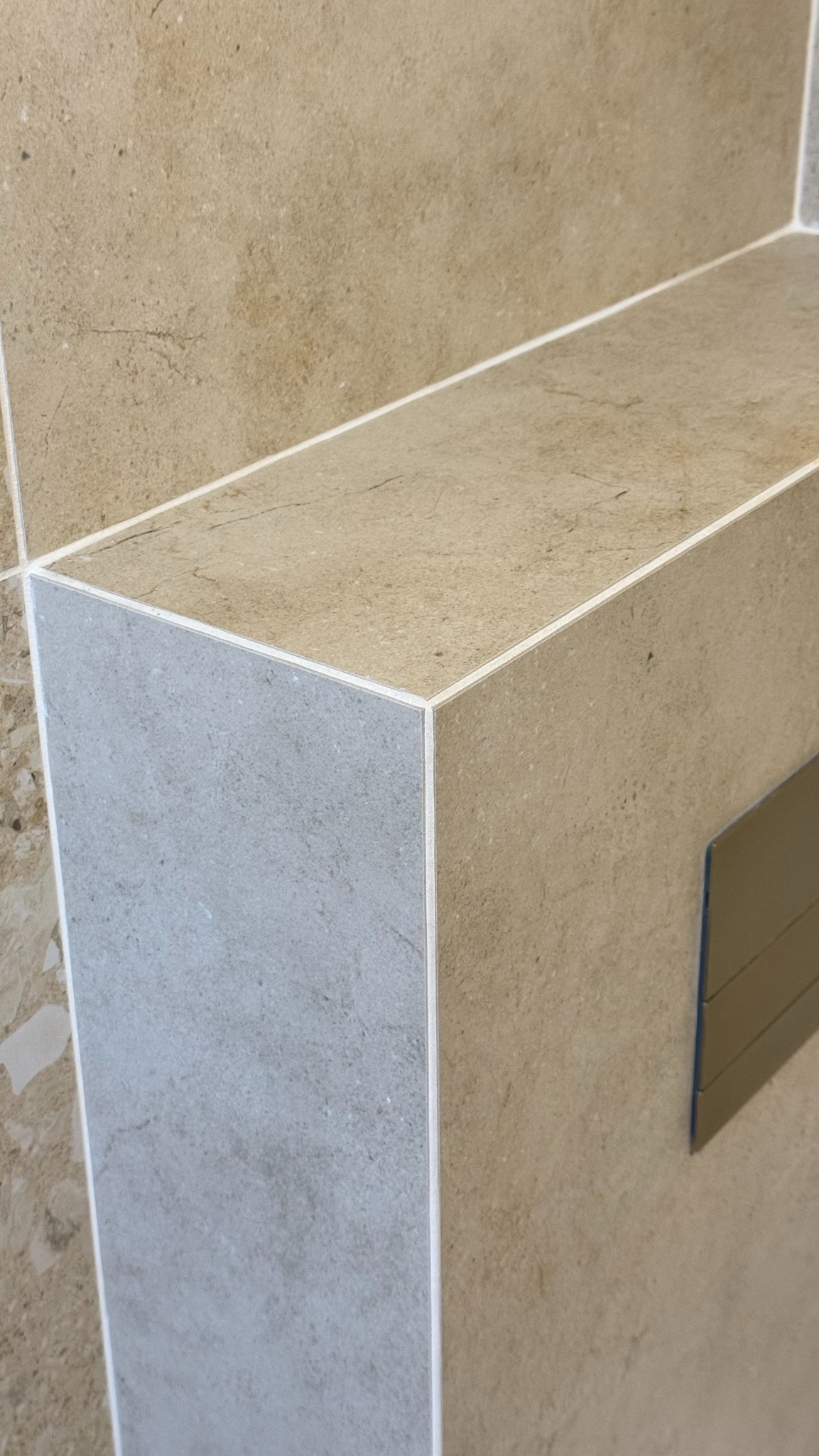 Wykończony fragment łazienki z beżowo-szarymi płytkami, widoczne fugi i przycisk spłuczki. Detal wykończenia narożnika z dbałością o szczegóły.