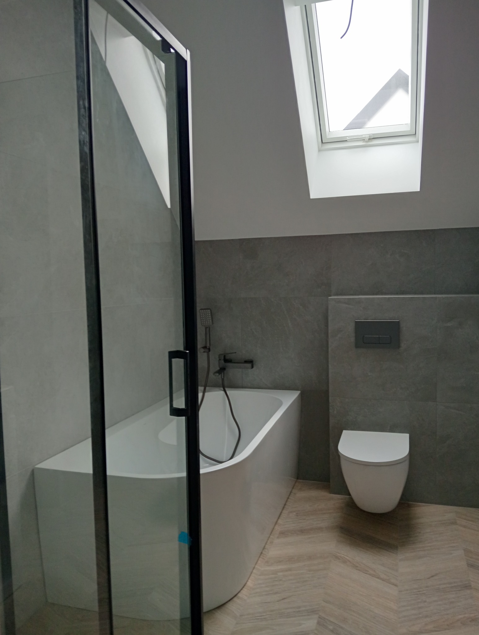 Nowoczesna łazienka na poddaszu z wanną za szklaną kabiną, szarą glazurą i oknem dachowym. Uwagę zwraca podłoga imitująca drewno ułożona w jodełkę.