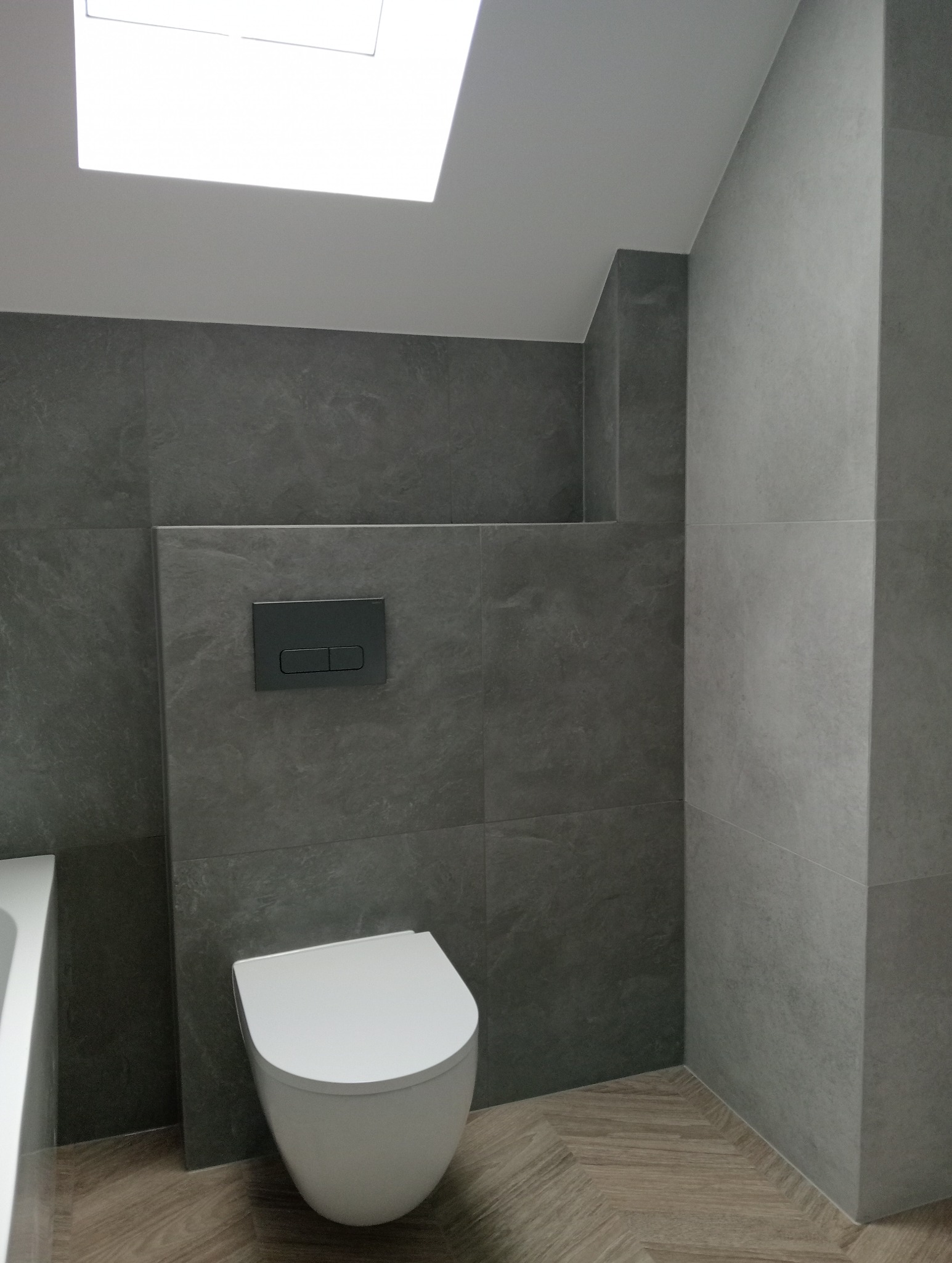 Nowoczesna łazienka z szarymi płytkami imitującymi kamień, podwieszana toaleta i drewnopodobna podłoga ułożona w jodełkę. Widoczne okno dachowe.