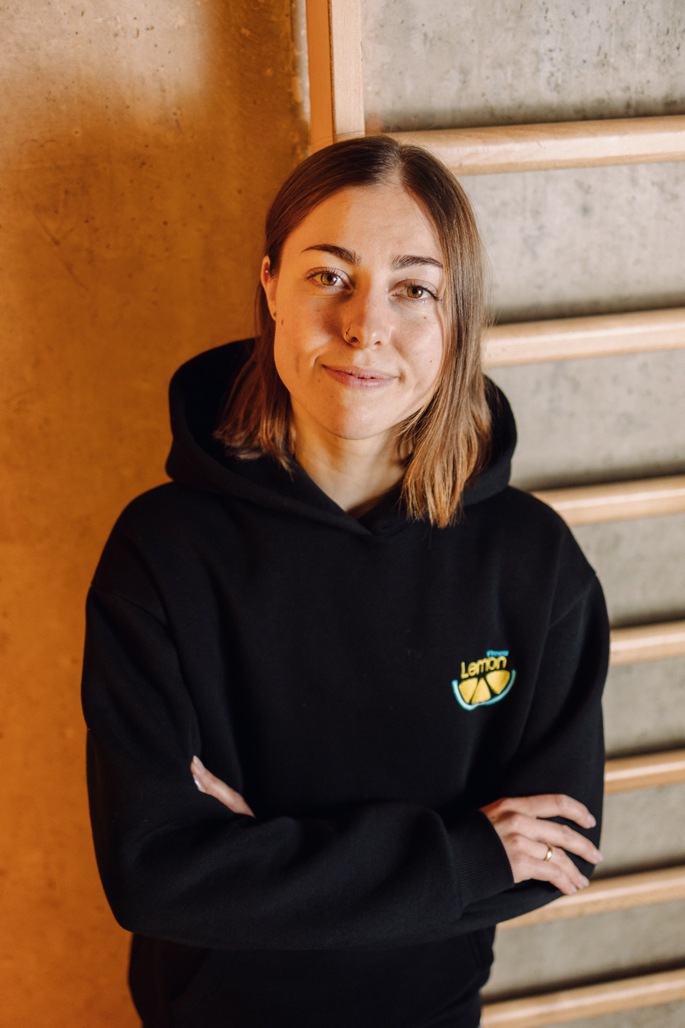 Portret kobiety w czarnej bluzie z logo Lemon, stojącej na tle drabinek gimnastycznych i betonowej ściany. Uśmiechnięta, z rękami skrzyżowanymi.