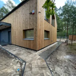 Nowoczesny dom z drewnianą elewacją i ścieżką z kostki brukowej. Ujęcie z poziomu gruntu, widoczne detale wykończenia i otoczenie domu.